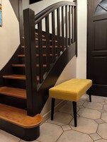 Uma escadaria com degraus de madeira e corrimãos pretos, um banco amarelo e uma porta ao fundo.