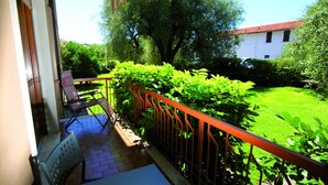 A balcony with a red railing, a chair, and a table. There is a lush green garden with various plants and a white building in the background.