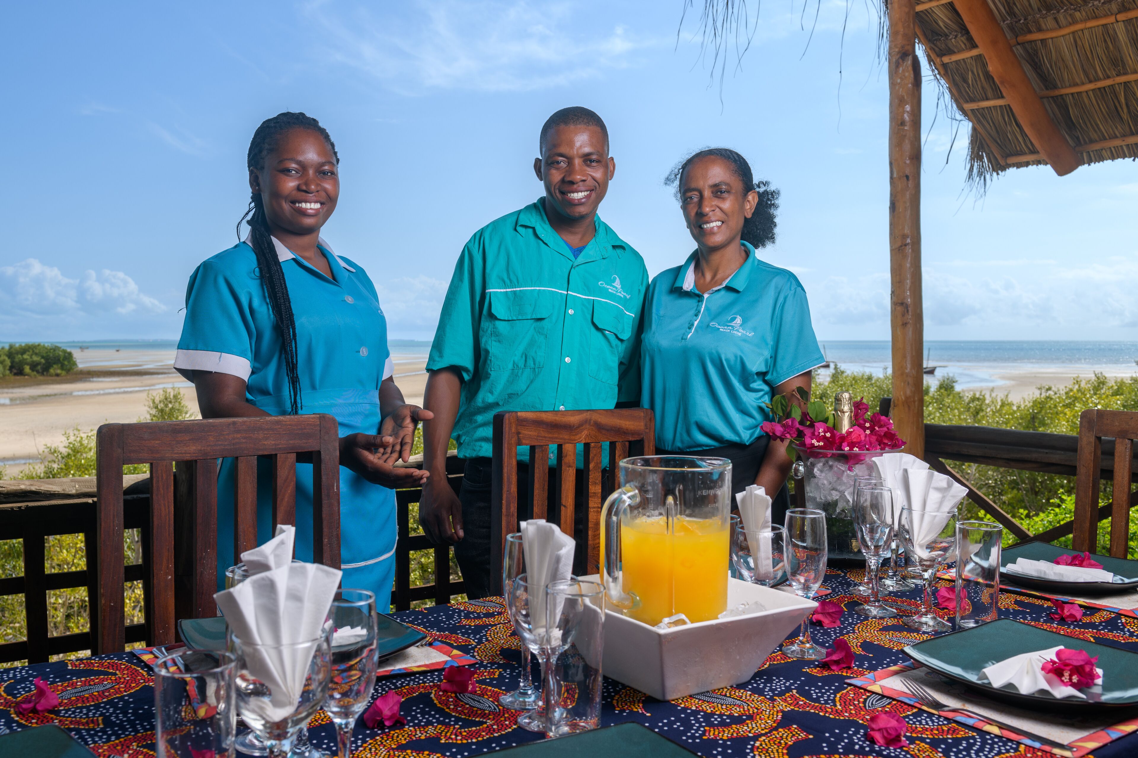 Three people seated at a table with a view of the beach, a table set with a pitcher of orange juice, and a thatched roof structure.