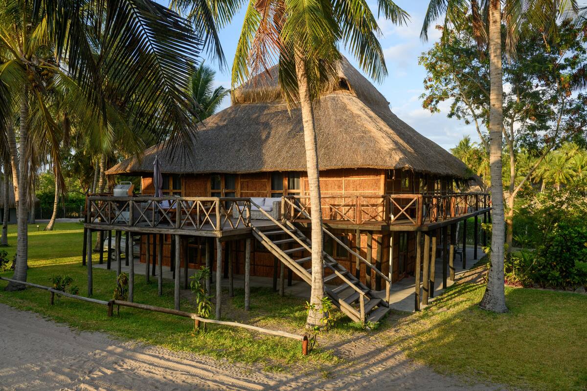 A thatched-roof bungalow with a wooden deck and staircase, surrounded by palm trees and greenery.