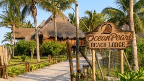 A wooden sign for Ocean Pearl Beach Lodge with a thatched-roof building in the background. - Ocean Pearl Beach Lodge (Vilanculos)