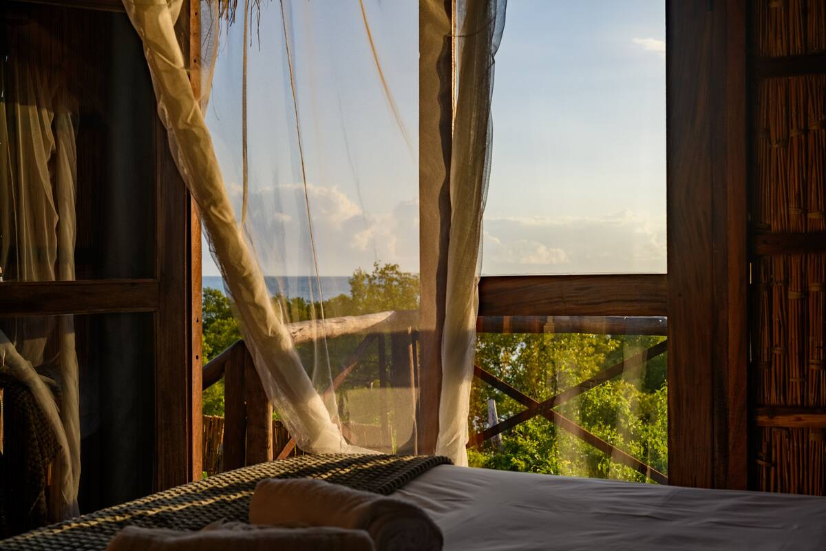 A room with a bed, a window with a view of greenery, and a wooden structure.
