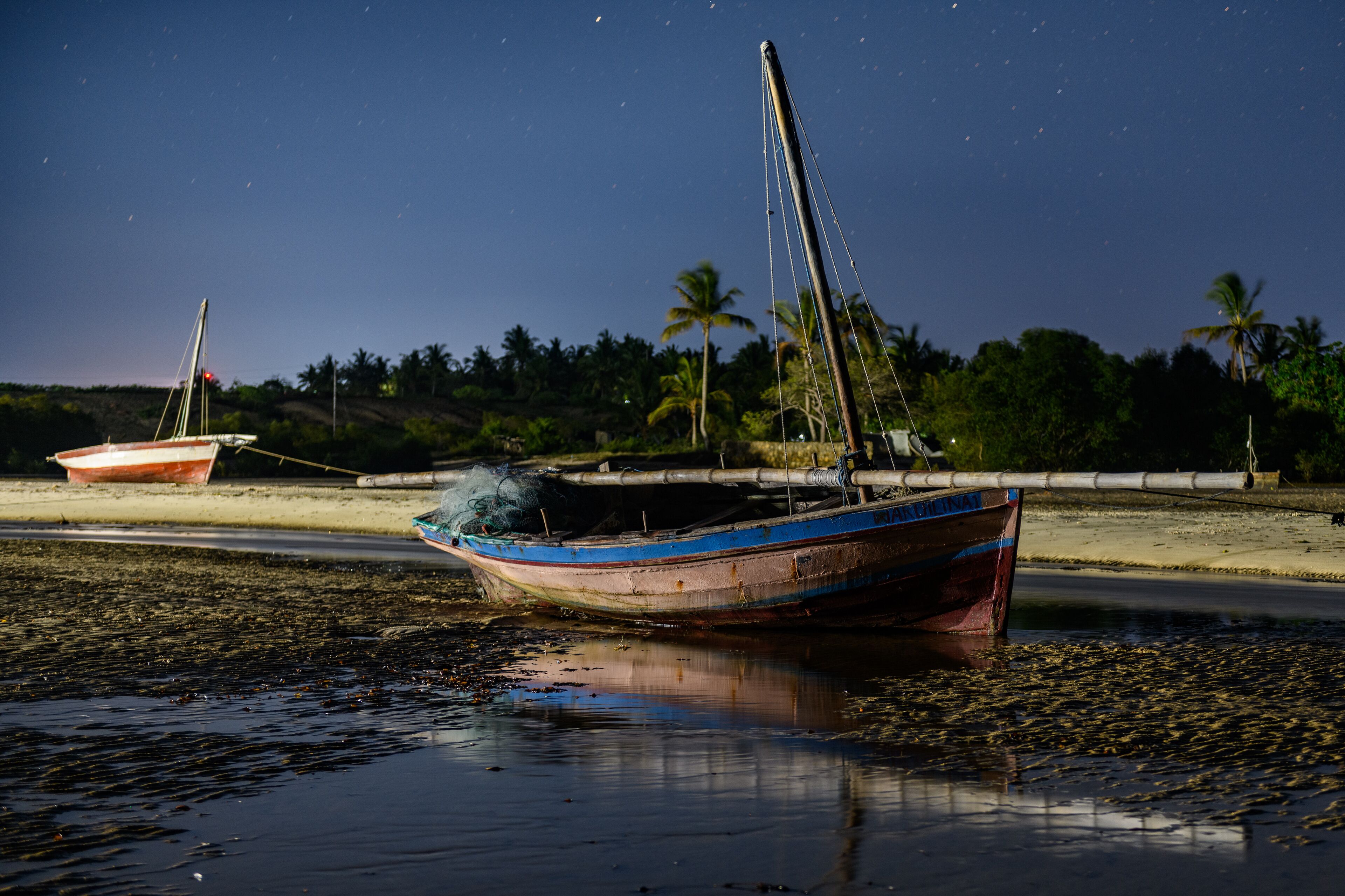 A boat tied to a dock with a sandy beach and palm trees in the background.