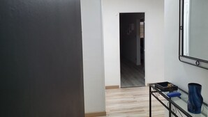En hall med en mörk vägg på ena sidan, en vit vägg på den andra och en dörr som leder till ett annat rum. Där står ett litet bord med en blå vas och ett svart bricka med diverse föremål på.