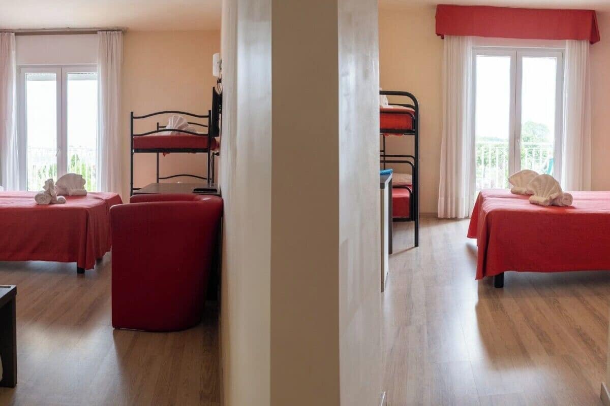 A hotel room with two single beds, a red armchair, a small table, and a balcony with a railing.