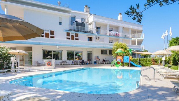 A swimming pool with lounge chairs and umbrellas, a children's play area, and a building with balconies. - Family Hotel Alexander (Jesolo)