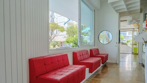 A reception area with a row of red cushioned seating, a large window with a view of buildings and trees, and a circular mirror on the wall. - Family Hotel Alexander (Jesolo)