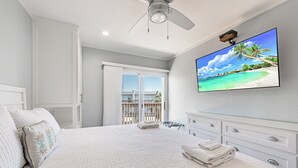 A bedroom with a large bed, a ceiling fan, a TV on the wall, and a view of the beach through the window.