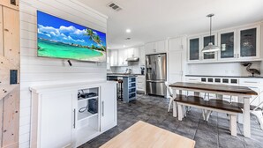 A modern kitchen with white cabinetry, a large flat-screen TV, a stainless steel refrigerator, and a wooden dining table with benches.