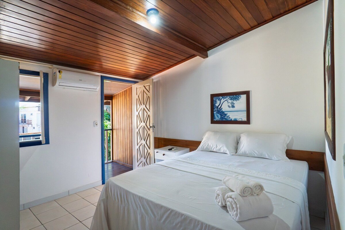 A bedroom with a wooden ceiling, a bed with white linens, a painting on the wall, and a door leading to an outdoor area.
