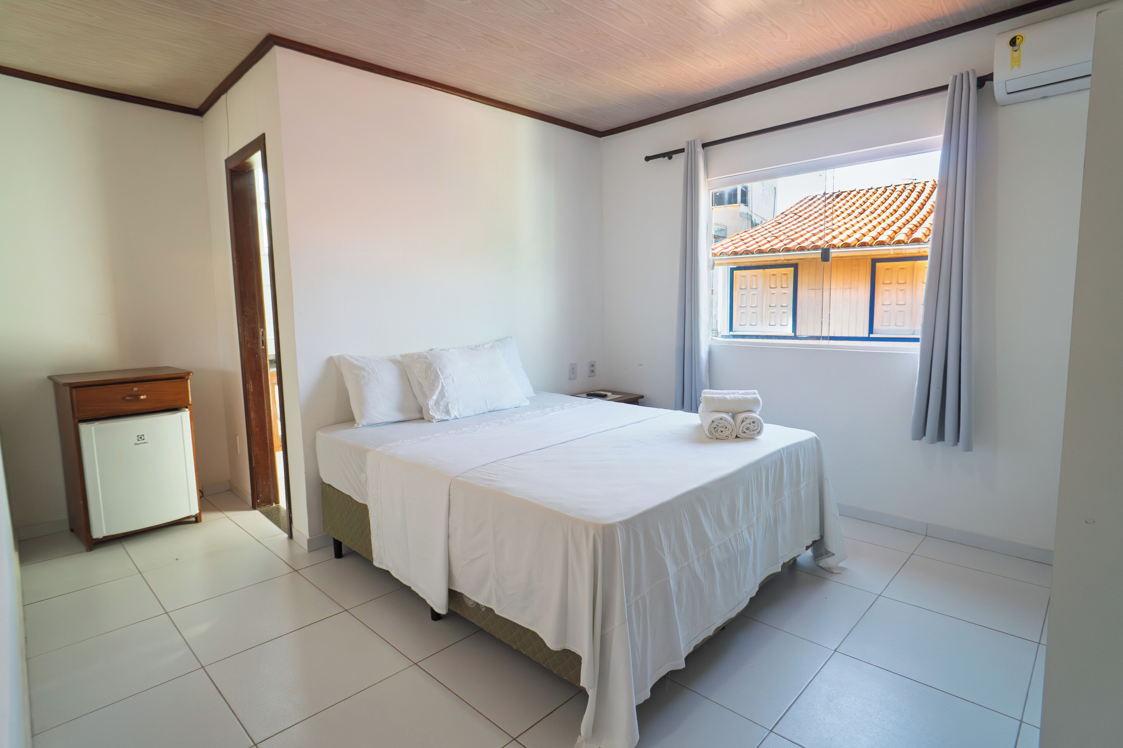 A bedroom with a bed, a nightstand, a small dresser, and a window with a view of a building with a tiled roof.