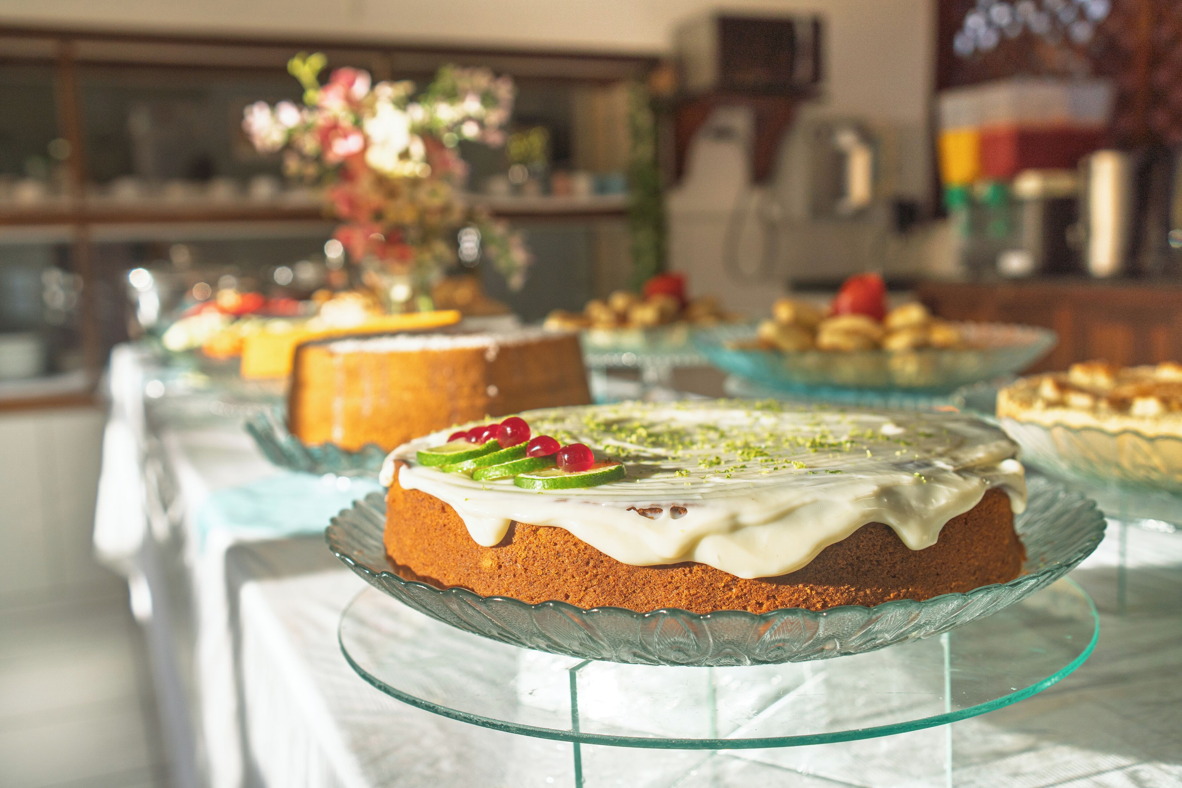 A cake on a glass stand with a white icing and green decorations, surrounded by other desserts on a table.