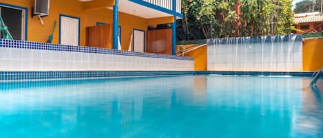 A swimming pool with a tiled edge, a balcony with blue railings, and a building with yellow walls and a tiled roof.