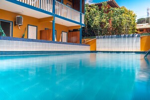 En swimmingpool med flisekant, en balkon med blå gelændere og en bygning med gule vægge og et tag med fliser.