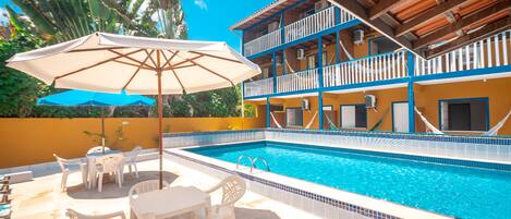 A swimming pool with a tiled edge, a covered seating area with chairs and tables, and a building with balconies.