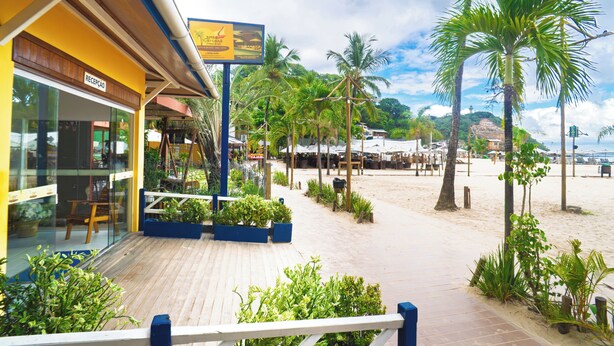 A wooden deck with potted plants, a reception area, and a beach view with palm trees. - Brisa do Caita Praia Hotel (Cairu)
