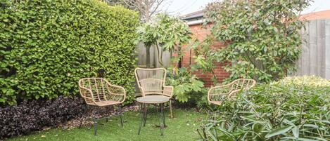 Un jardin avec trois chaises en osier, une petite table ronde et diverses plantes.