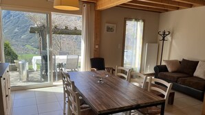 A dining area with a wooden table and chairs, a sofa, and a view of mountains through large windows. - Maison à Camparan Proche de Saint-lary (Camparan)