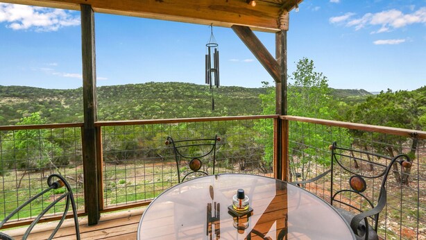 Un terrazzo in legno con tavolo in vetro e sedie in metallo, affacciato su un'area recintata con alberi e colline.
