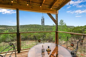 Una terraza de madera con una mesa de vidrio y sillas de metal, con vista a un área cercada con árboles y colinas.
