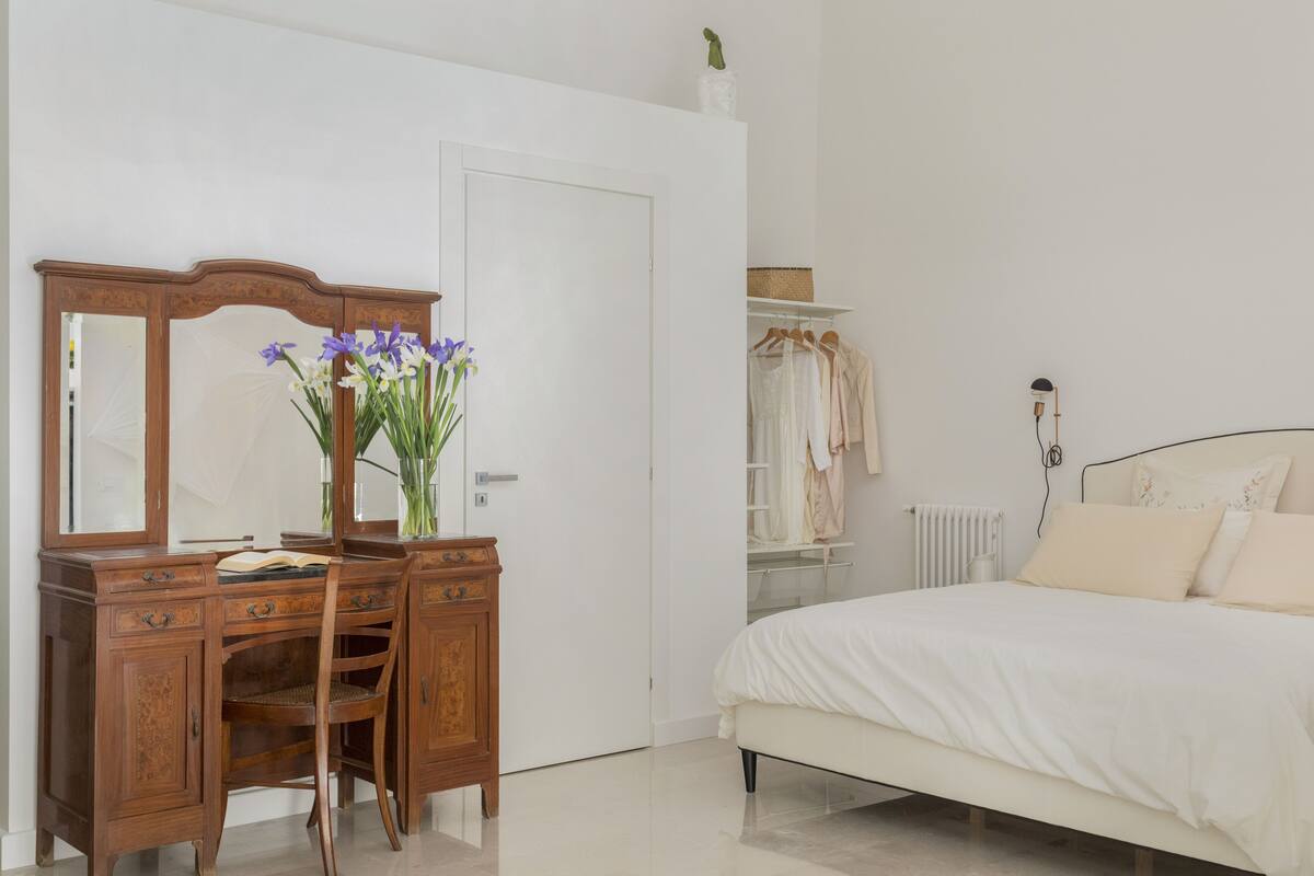 Une chambre à coucher avec une commode en bois, un lit avec une literie blanche, un miroir et un vase de fleurs.