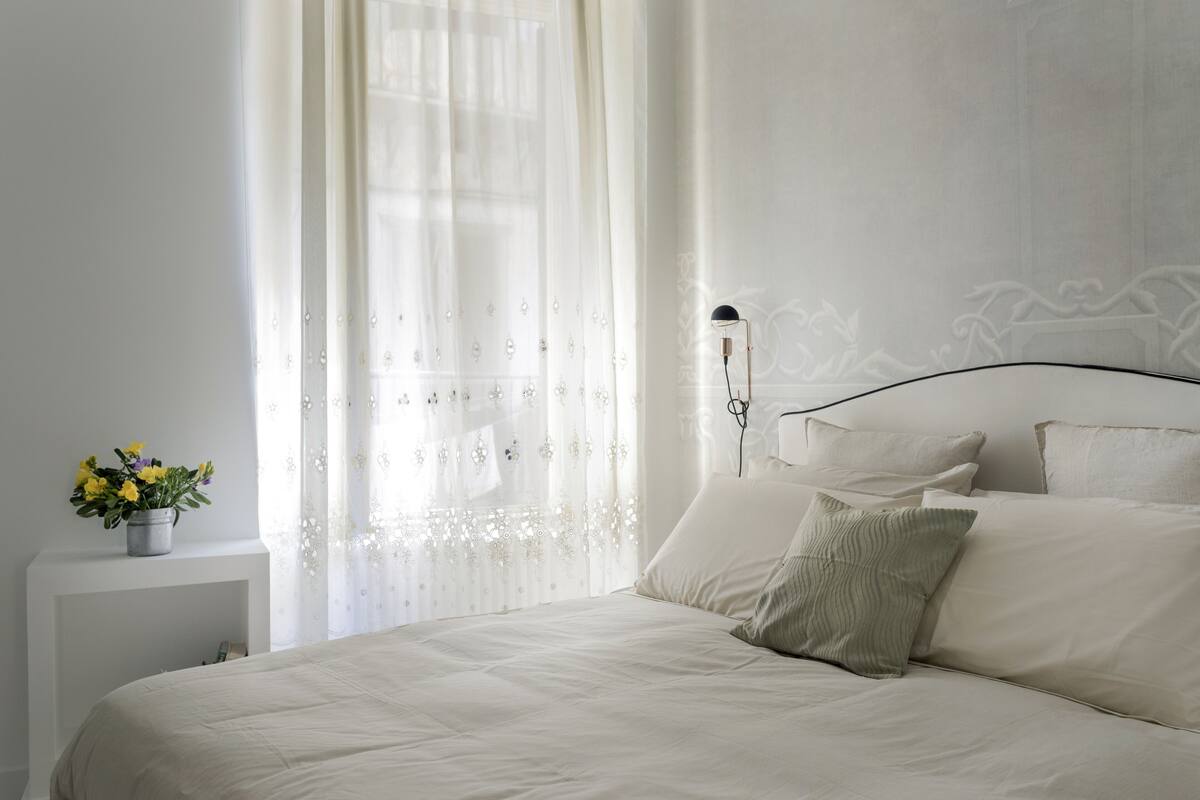 Un lit bien fait, avec une literie blanche, une table de chevet avec une lampe et des rideaux légers ornés de motifs floraux.