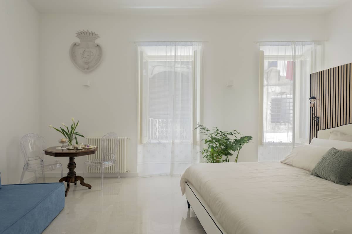 Une chambre à coucher avec un lit, une petite table sur laquelle est posée une plante, un radiateur et deux fenêtres ornées de rideaux légers.