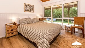 Un dormitorio con una cama grande, una mesita de noche con lámpara, muebles de mimbre y vistas al verdor a través de amplias ventanas.