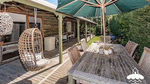 Una terraza de madera con una mesa y sillas, una silla colgante de mimbre y un paraguas verde.