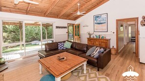 A spacious living room with a brown leather sofa, a wooden coffee table, and a large window offering a view of the outdoors.