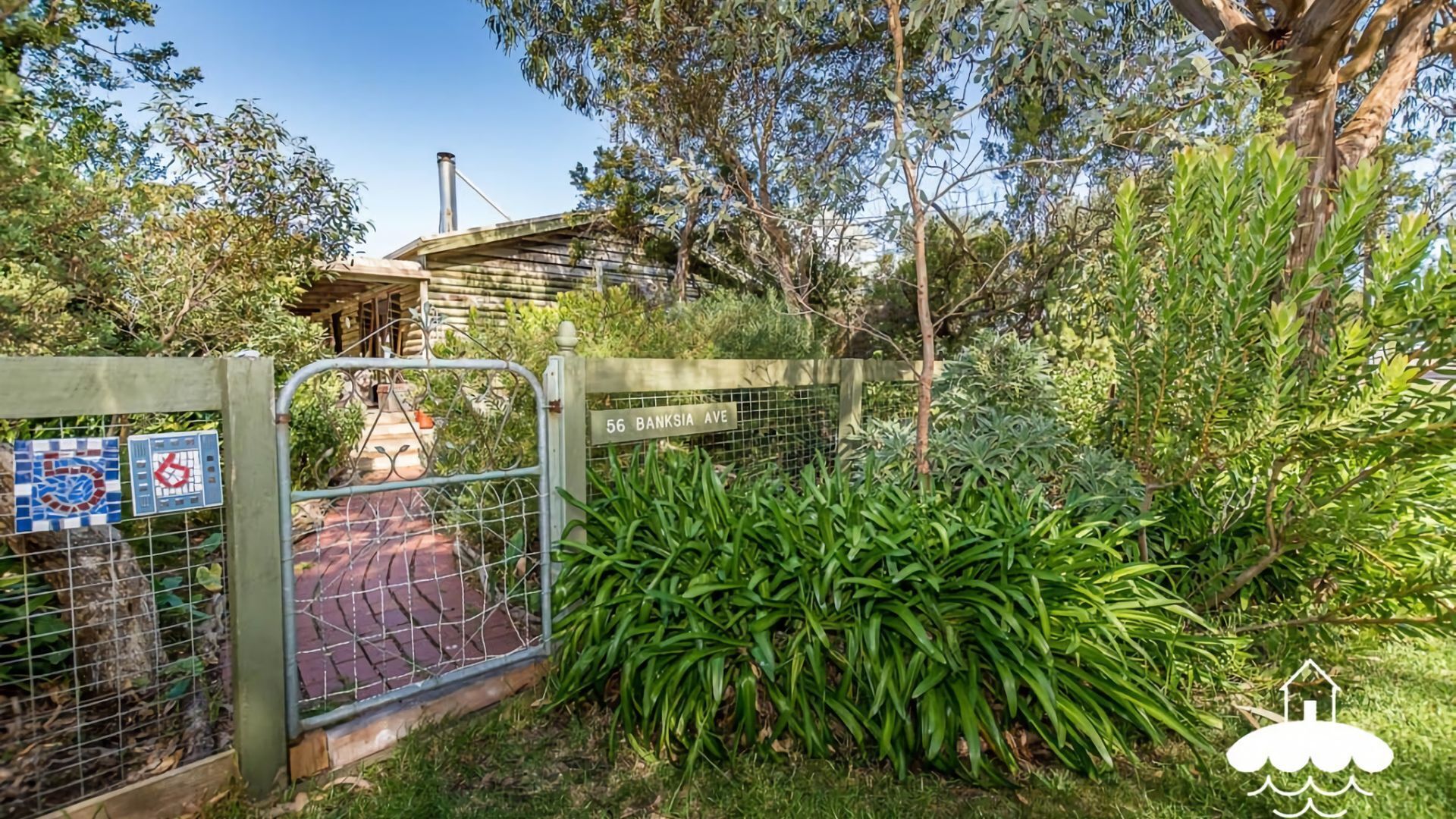 A gated entrance with a metal gate, a sign reading "56 Banksia Ave", and a pathway leading to a building with a chimney.
