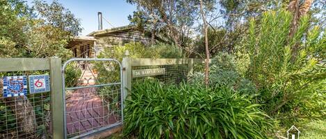 A gated entrance with a metal gate, a sign reading "56 Banksia Ave", and a pathway leading to a building with a chimney.