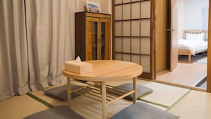 Une pièce de style japonais traditionnel, avec un sol recouvert de tatamis, une petite table en bois avec un coussin, un meuble orné d’un tableau et une porte coulissante donnant sur un lit.