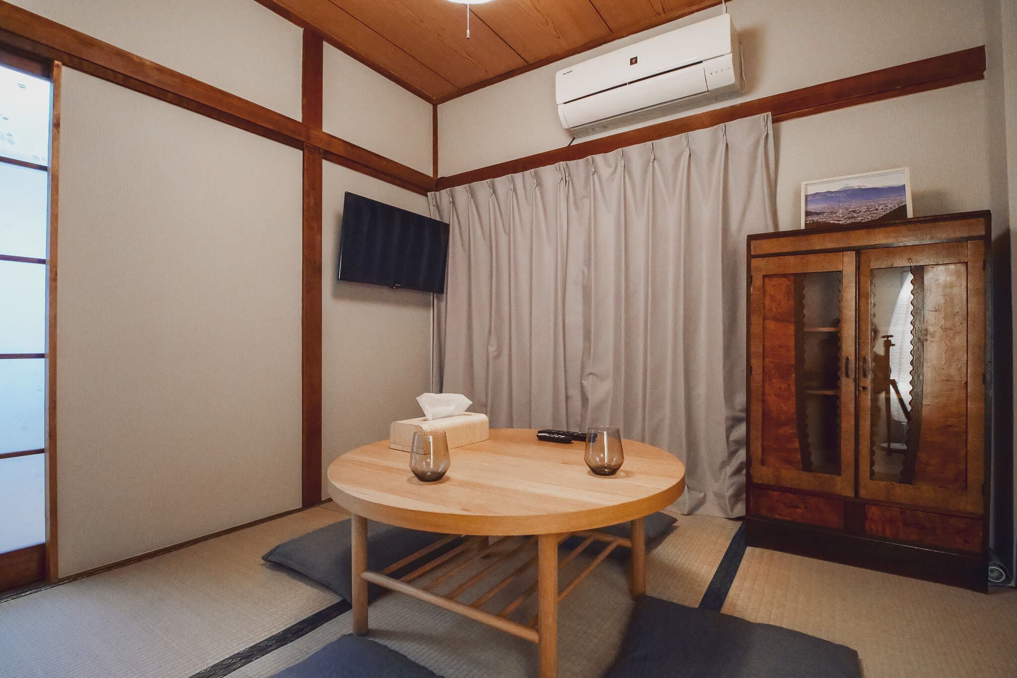 A traditional Japanese-style room with tatami flooring, a wooden table with two glasses, a remote control, and a cabinet with a picture on top.