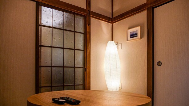 Une pièce de style japonais traditionnel, avec un sol recouvert de tatamis, un paravent shoji, une table ronde en bois équipée d’une télécommande et un luminaire fixé au mur.