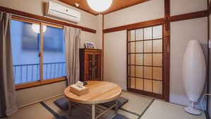 Une pièce de style japonais traditionnel, avec un sol recouvert de tatamis, une table ronde en bois, un futon servant de lit, des portes coulissantes et un ventilateur de plafond.