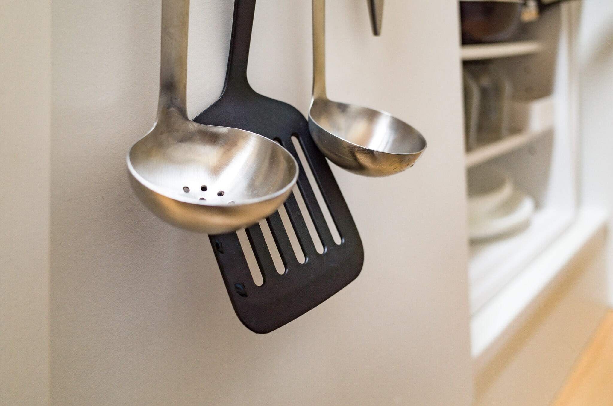 A kitchen with a white wall, a black spatula, and a stainless steel ladle hanging on the wall.