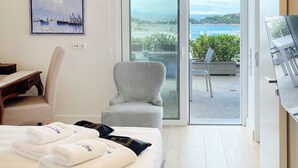 A hotel room with a bed, a chair, a desk, a TV, and a balcony with a view of the sea. - A unique 3 level penthouse with a great sea view to Vouliagmeni Bay (VOULIAGMENI)