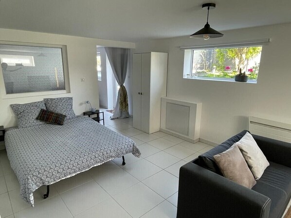 A bedroom with a bed, a sofa, a window with a view of greenery, a radiator, and a ceiling light.