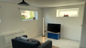 Un salon moderne avec un canapé noir, un téléviseur à écran plat posé sur un meuble bleu et une fenêtre donnant sur un paysage verdoyant.