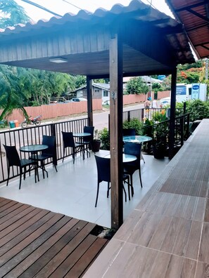 An outdoor patio with tables and chairs, a wooden deck, and a view of the street and greenery.