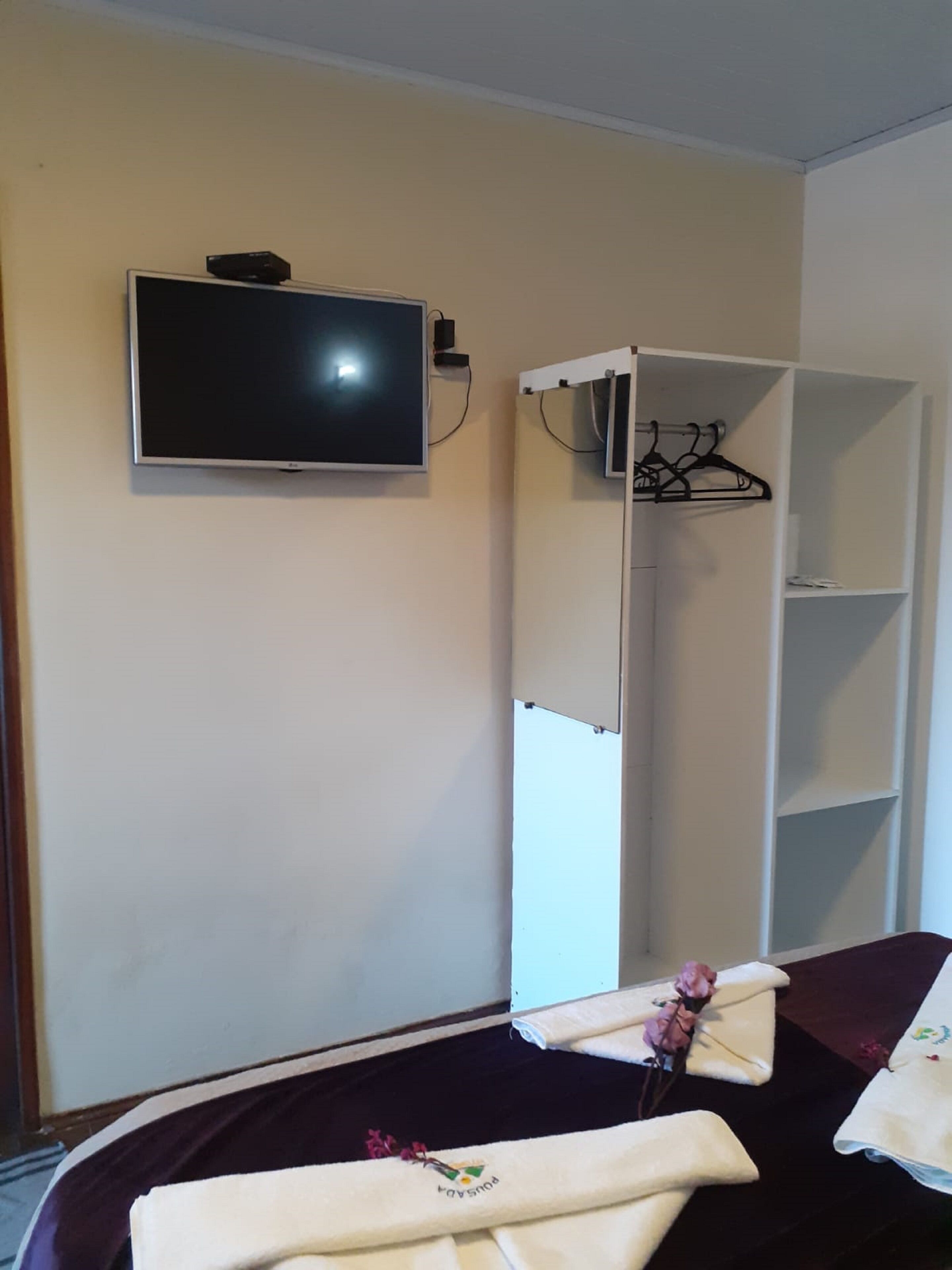 Une chambre avec un lit, un téléviseur fixé au mur, une armoire et une petite table sur laquelle se trouvent une serviette et des fleurs.