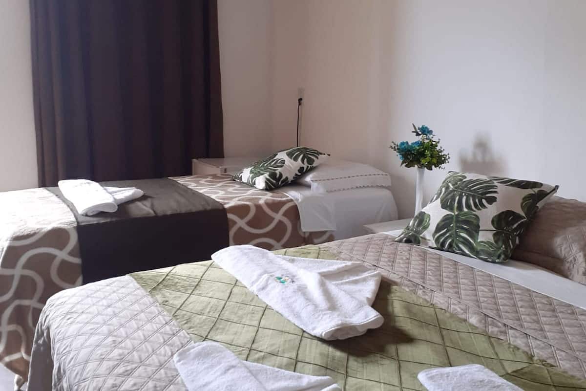 Quarto de hotel com duas camas, cada uma com colcha acolchoada e um travesseiro. Há uma janela com cortinas, uma mesa pequena e um vaso com flores.
