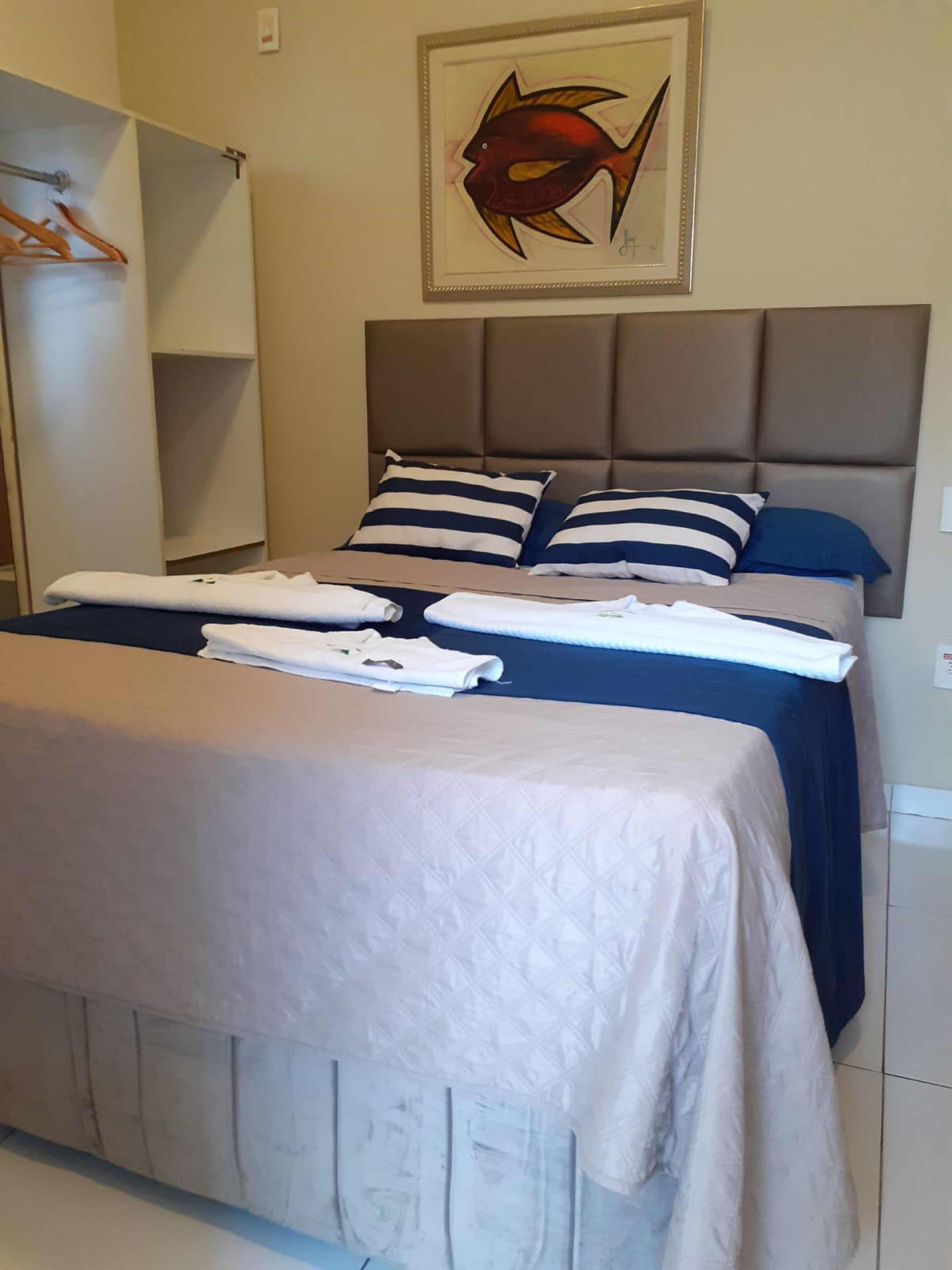 Un lit bien fait, avec des oreillers à rayures et une tête de lit, une serviette posée sur le lit et, au mur, un tableau représentant un poisson, encadré.