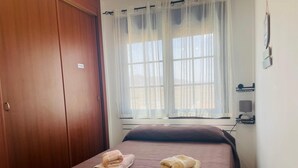 Une chambre à coucher avec un lit, une porte en bois, une fenêtre avec des rideaux légers, une table de chevet avec une lampe et une étagère fixée au mur.