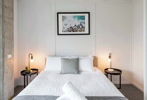 Een net opgemaakt bed met een witte deken en een grijze kussen, twee nachtkastjes met lampen, een ingelijste foto aan de muur, en een betonnen muur aan de linkerkant.
