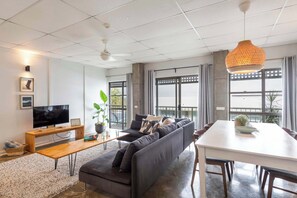 Een moderne woonkamer met een bank, een televisie, een eettafel en grote ramen.