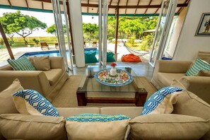 A spacious living area with a glass coffee table, beige sofas, and patterned cushions. A view of a pool and outdoor seating is visible through the glass. - Kay Jay Beach House - Pasikudha (Pasikuda)