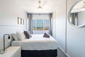A hotel room with a bed, a bedside table, a lamp, a window with curtains, and a round mirror on the wall. - Yamba Views Iluka Apartment (Yamba)