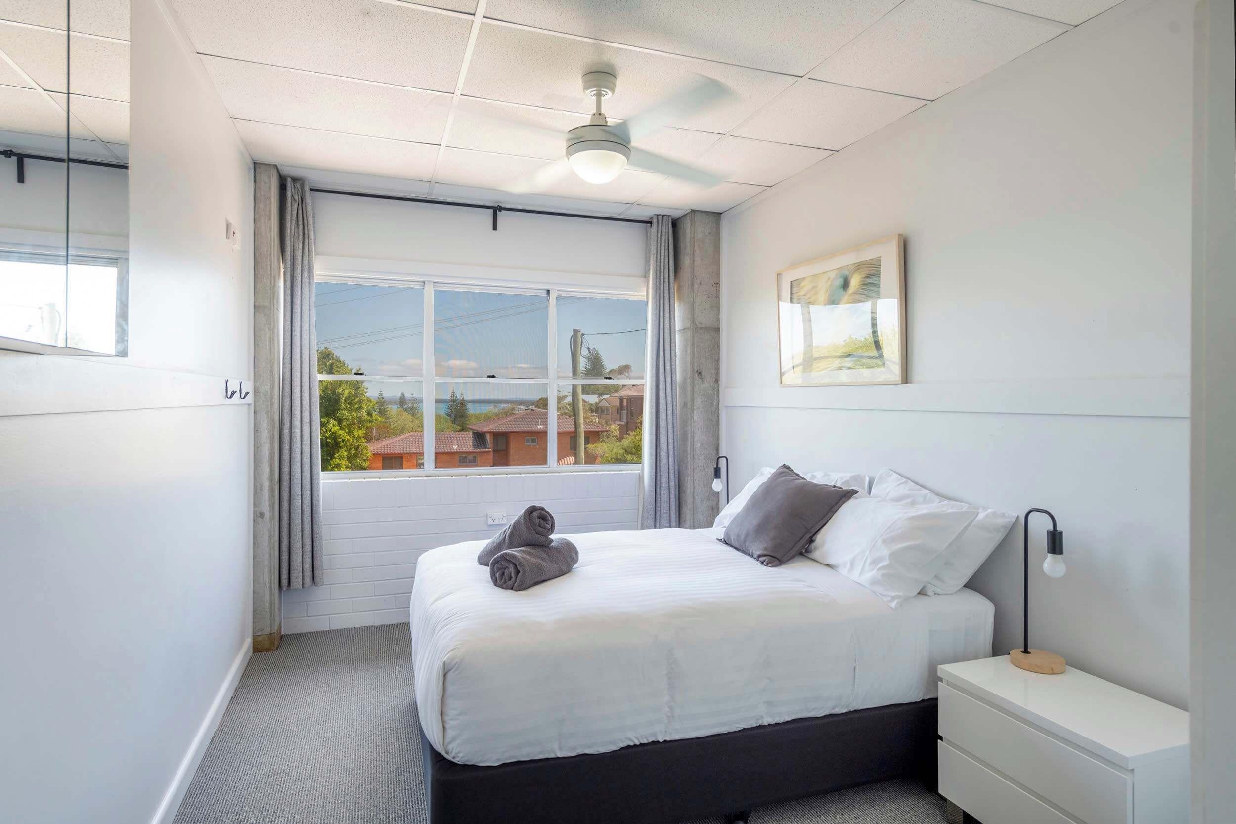 A bedroom with a bed, a bedside table, a ceiling fan, a window with curtains, and a framed picture on the wall.
